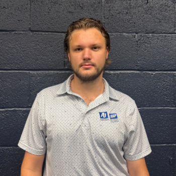kyle thomas edited headshot | AJ Adhesives Male adult with blond hair and stubble stands against a dark brick wall, wearing a light gray polo with a blue AJMAP logo on the chest.