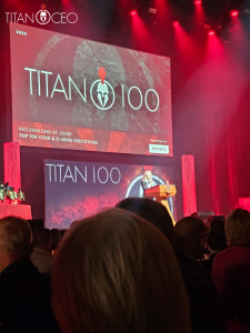 Titan 100 Event 5 | AJ Adhesives Stage at Titan 100 awards event with a presenter at a wooden podium, a large screen reading TITAN 100 and red lighting, audience in foreground.