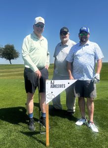 Bill McDonnell - Golf Tournament Sponsor | AJ Adhesives Three men posing on a sunny golf course beside a triangular sponsor sign reading 'AJ ADHESIVES' on a wooden stake.